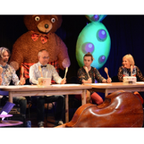 On stage, four actors are sitting at a table. They hold raised wooden spoons.
