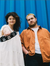 Photo of the band members: a woman in a white dress and a man in an orange jacket. Blue background.