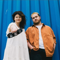 Photo of the band members: a woman in a white dress and a man in an orange jacket. Blue background.