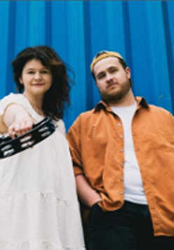 Photo of the band members: a woman in a white dress and a man in an orange jacket. Blue background.
