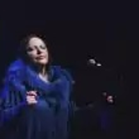 Photo of Grażyna Łobaszewska standing by a microphone, dark background.