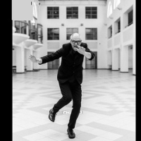 Black and wjite photo of the artist. Mariusz Lubomski on the move, in a black suit and glasses on his nose. He poses in front of a large, bright corridor
