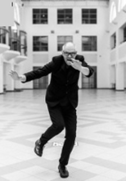 Black and wjite photo of the artist. Mariusz Lubomski on the move, in a black suit and glasses on his nose. He poses in front of a large, bright corridor