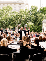 Photo of a orchestra performing outside. The conductor in front of the orchestra, the audience behind him, green trees as a background.