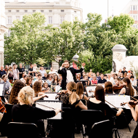Photo of a orchestra performing outside. The conductor in front of the orchestra, the audience behind him, green trees as a background.