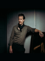 A man stands in a relaxed pose, looking straight ahead, leaning with one hand on a piece of furniture.