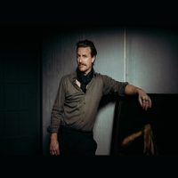 A man stands in a relaxed pose, looking straight ahead, leaning with one hand on a piece of furniture.