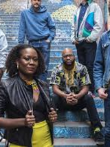 Photo of the band: one woman and five man on the outdoor stairs.