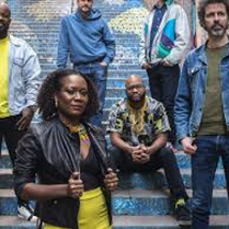 Photo of the band: one woman and five man on the outdoor stairs.