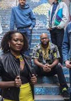 Photo of the band: one woman and five man on the outdoor stairs.