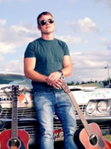 Photo of Rodney Branigan - a young man sitting on a car's bonnet, with two guitars on his left and right.
