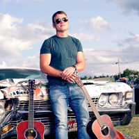 Photo of Rodney Branigan - a young man sitting on a car's bonnet, with two guitars on his left and right.