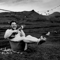 Black and white outside-taken photo of a man lying in a small tub and playing the guitar.