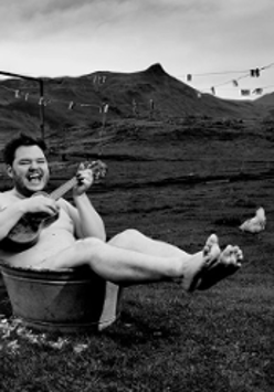 Black and white outside-taken photo of a man lying in a small tub and playing the guitar.