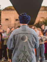 Photo of a group of singing people. In the foreground, one person with his back turned back. Green bushes and trees, and a building as a background.