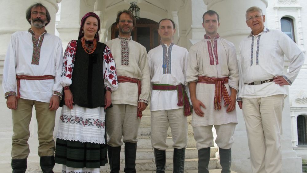 Photo of the band - 6 men and a woman in traditional folk costumes. A white building as a background.