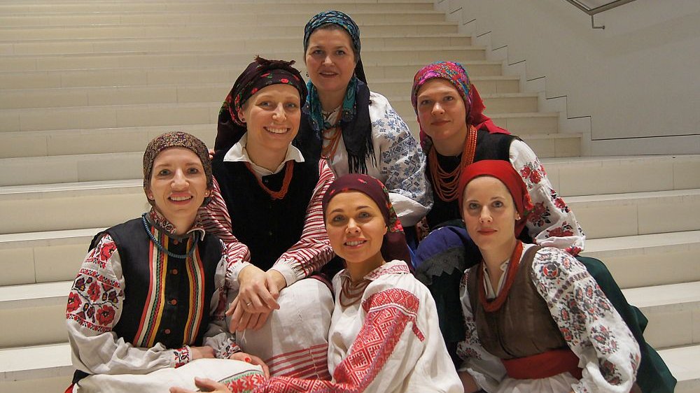 Photo of the band - six women in traditional folk costumes on white stairs.