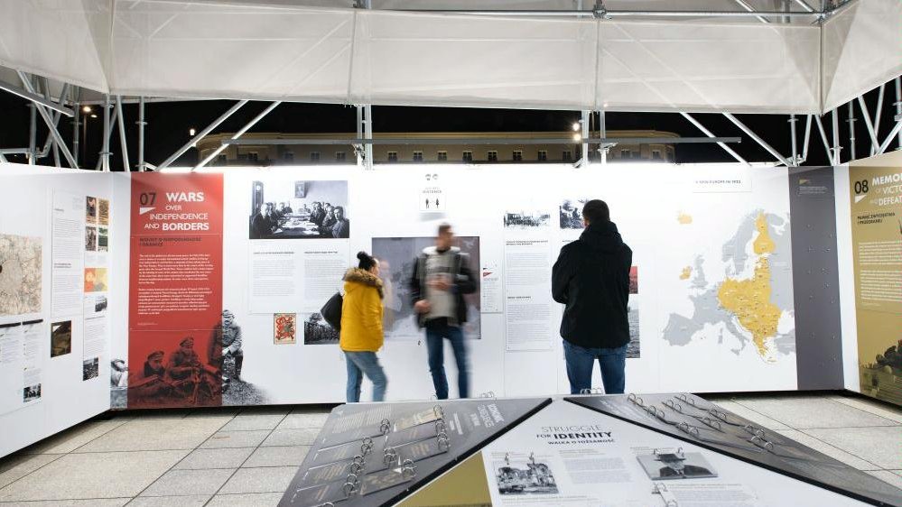 Photo of three people watching the exhibition. On the walls and in the middle of the presented space pictures and descriptions from the exposition.