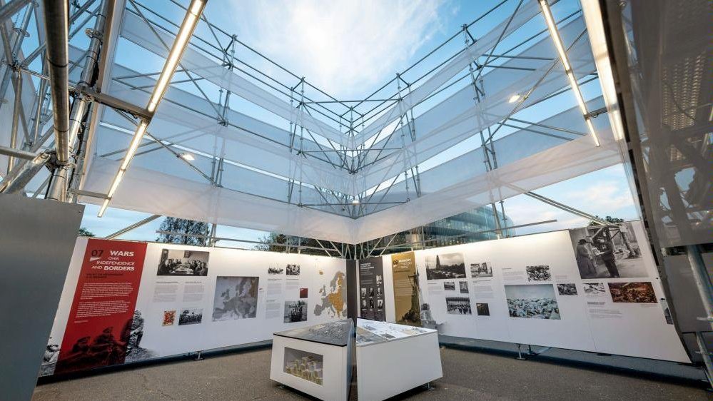 Exhibition interior: white walls with photos and descriptions, in the center a stand with several photos and descriptions. As the exhibition is presented in a metal openwork structure, a fragment of the building from the outside and the sky are visible.
