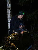 In the photo there are two men in the forest. One of them is wrapped in a gold and silver thermal emergency blanket and has her eyes closed. The other, standing above him with a headlamp on his head, holds him.