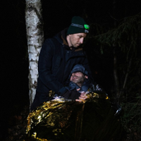 In the photo there are two men in the forest. One of them is wrapped in a gold and silver thermal emergency blanket and has her eyes closed. The other, standing above him with a headlamp on his head, holds him.