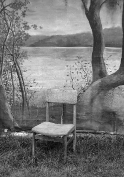 Black and white photo shows a chair standing on the ground covered with grass. Behind the chair there is an old photographic background with painted trees, water and blurry mountains on the horizon.