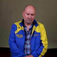 Exhibition poster: photo of a sad looking man in blue and yellow shirt and jacket.