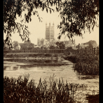 Sepia photograph of the Gloucester Cathedral, which is in the background. In the foreground a lake or a river with some water plants.
