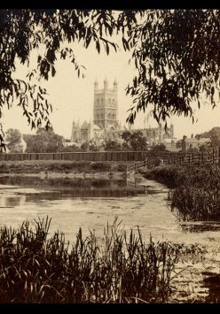 Sepia photograph of the Gloucester Cathedral, which is in the background. In the foreground a lake or a river with some water plants.