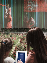 Two girls, standing back to the camera, in front of a mockup made of Lego bricks.
