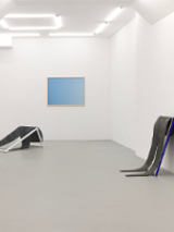 Picture of the white room with one blue picture on the wall and two exhibits by the walls.