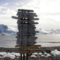 One of the exhibition works: a photo of a pole with many signposts with names of places and distances to them. In the background water and snowy mountains.