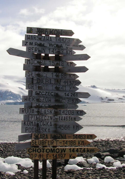 One of the exhibition works: a photo of a pole with many signposts with names of places and distances to them. In the background water and snowy mountains.