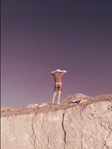 A picture of half-naked man standing on a rock, wearing white knee socks, long white gloves, white cap and white thong.