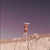A picture of half-naked man standing on a rock, wearing white knee socks, long white gloves, white cap and white thong.