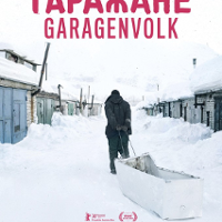 A man pulling a white container-sled in the snow. Low buildings covered with snow around.