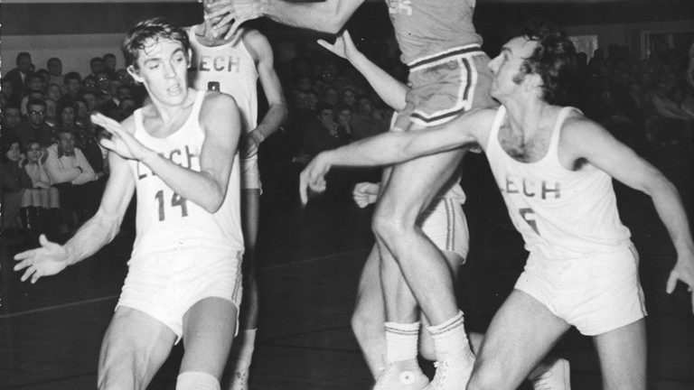 Black and white photo of five men playing basketball. One of them in a jump.