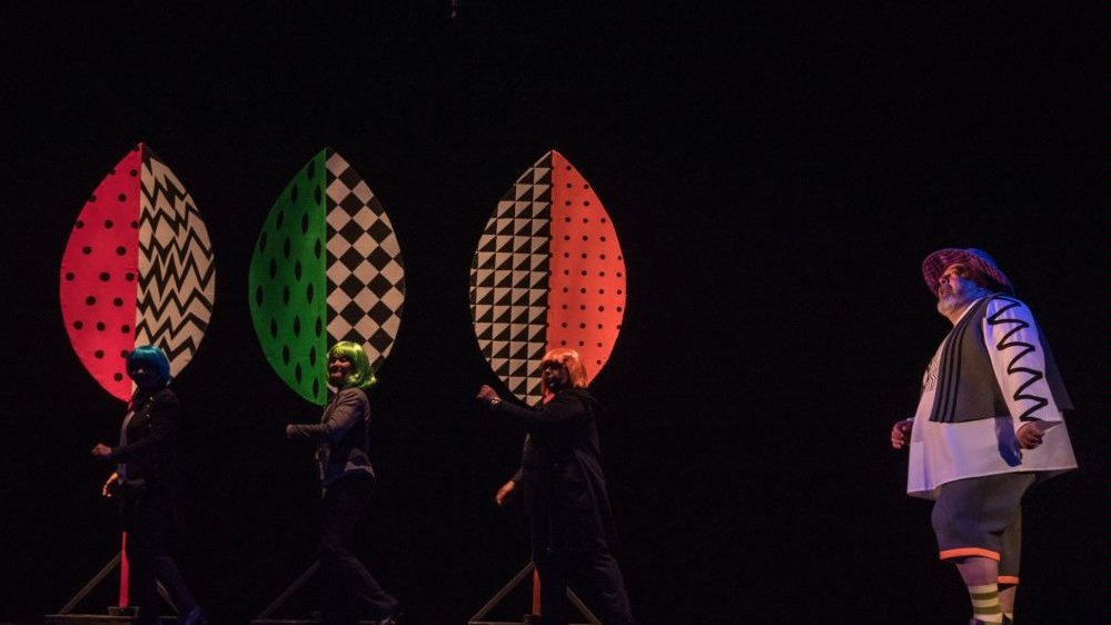 Photo from the performance - four actors on stage and three colourful trees on a black background.