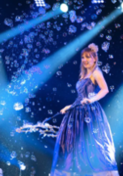 Photo from the performance - a woman in long, blue dress on stage blowing bubbles.