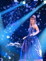 Photo from the performance - a woman in long, blue dress on stage blowing bubbles.