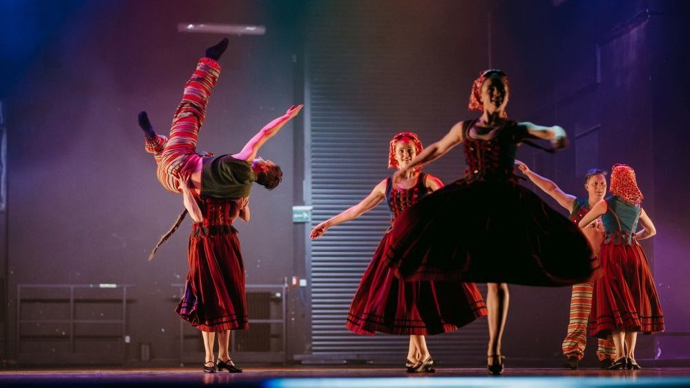 Photo from the show. On the right, dancers in dance poses in folk costumes on the stage. On the left, a woman holding a man.