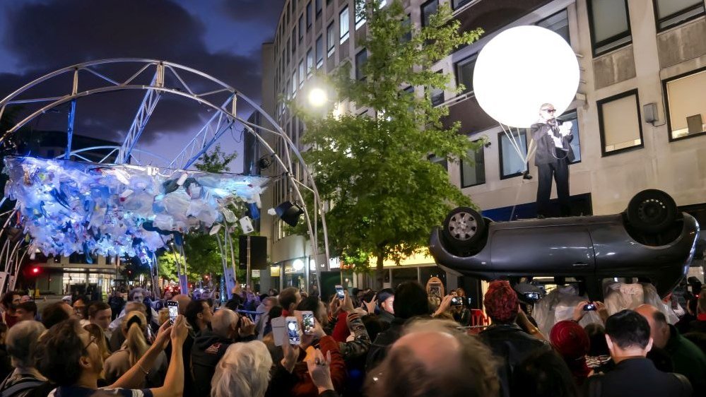 A scene from the performance - a man standing on a car which is upside down, behind him a big white balloon. In a foreground people watching a performance, in a background - a building and a tree.