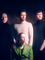 Photo of the band members - three men who are standing behind a woman who is sitting