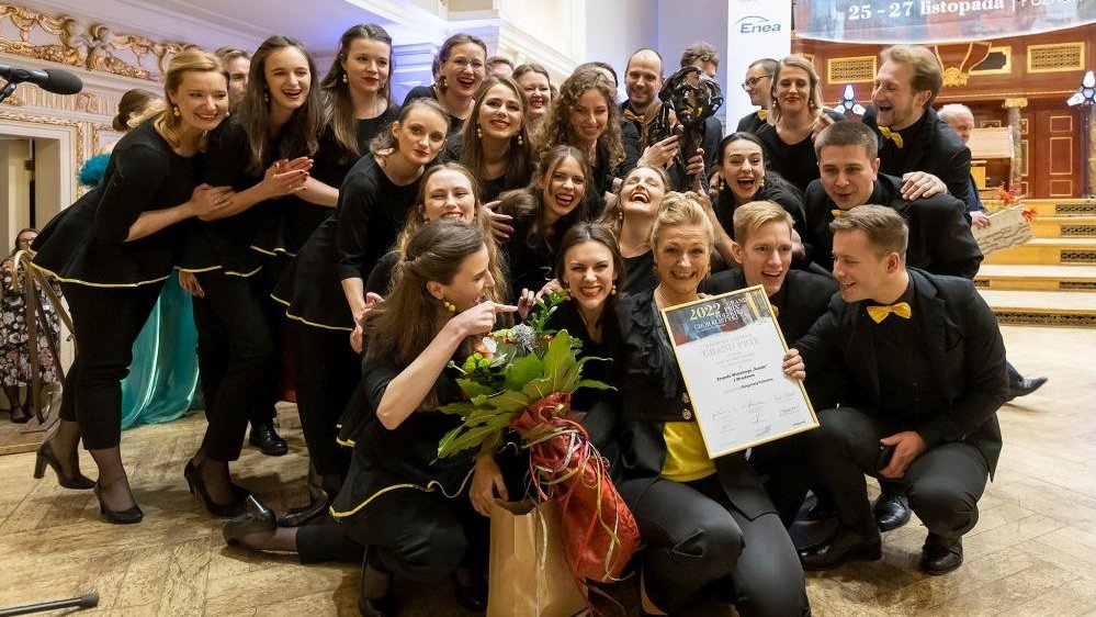Photo of the choir members, young women and men, standing or in a squat, smiling. One of the women is holding bunch of flowers, the other is holding a diploma.