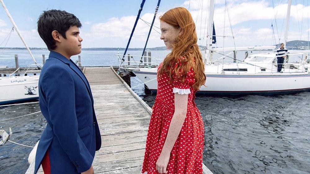 A picture of a red-haired girl and a boy who are looking at each other. The children are standing on a pier, a yacht and a lake in the background.