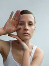The photo shows a portrait of the artist in a white T-shirt. Her hair smoothly combed back. She touches her face with her hands slightly stained with chalk.