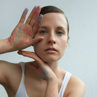 The photo shows a portrait of the artist in a white T-shirt. Her hair smoothly combed back. She touches her face with her hands slightly stained with chalk.