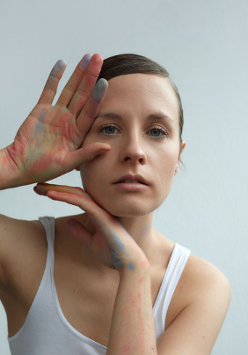 The photo shows a portrait of the artist in a white T-shirt. Her hair smoothly combed back. She touches her face with her hands slightly stained with chalk.