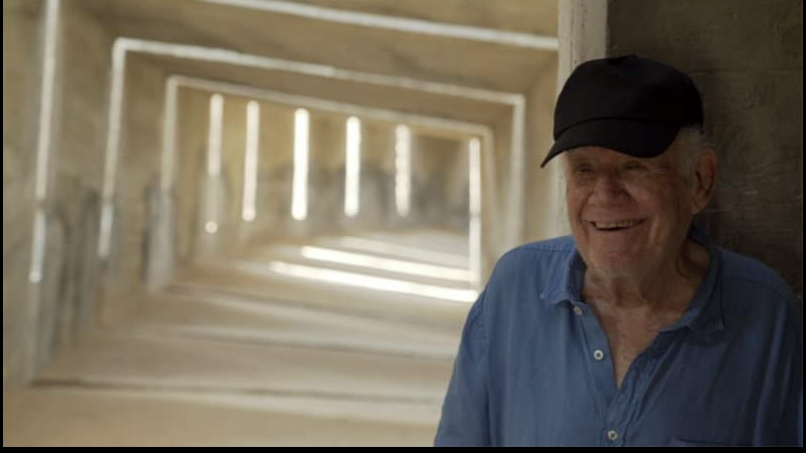 A photo from the movie - on the right an elderly man who is smiling and leaning against the wall. An empty concrete space as a background.