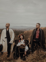 Photo of the band: a woman sitting and two men standing in folk clothes; mountainous landscape in the background